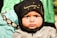 A young child with a thoughtful and serious expression, wearing a black headscarf with Arabic script. The child's jacket is gray with a shiny texture, and there is a background of green and black fabric.