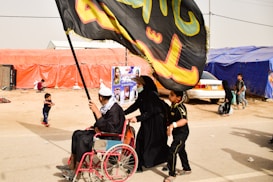 A person in a wheelchair is being pushed by another individual, both dressed in black. They are holding a large, black flag with colorful writing. A child walks alongside them. In the background, there are tents made of orange and blue fabric, a car, and other people, including a child running.