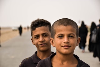 Two young boys stand close together along a pathway that stretches into the distance. The background is populated with several figures wearing dark clothing, and a few cars can be seen further along the path. The setting appears to be on a sunny day with a clear sky.