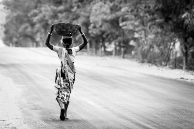 A person wearing traditional clothing walks down an empty road with a basket balanced on their head. The surrounding area is rural with trees lining the sides of the road, and the image is in black and white.
