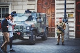 A camouflaged military vehicle is parked on a cobblestone street in front of a large wooden door with ornate designs. A soldier in uniform stands guard beside the vehicle holding a weapon. Two civilians are walking past, engaged in conversation.