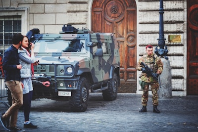A camouflaged military vehicle is parked on a cobblestone street in front of a large wooden door with ornate designs. A soldier in uniform stands guard beside the vehicle holding a weapon. Two civilians are walking past, engaged in conversation.