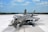 A modern military jet fighter is positioned on an expansive runway under a bright blue sky. The aircraft features sleek, aerodynamic lines and is painted in a gray camouflage pattern. It is equipped with various weapons and has a glossy cockpit canopy.