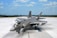 A modern military jet fighter is positioned on an expansive runway under a bright blue sky. The aircraft features sleek, aerodynamic lines and is painted in a gray camouflage pattern. It is equipped with various weapons and has a glossy cockpit canopy.