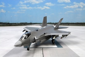 A modern military jet fighter is positioned on an expansive runway under a bright blue sky. The aircraft features sleek, aerodynamic lines and is painted in a gray camouflage pattern. It is equipped with various weapons and has a glossy cockpit canopy.