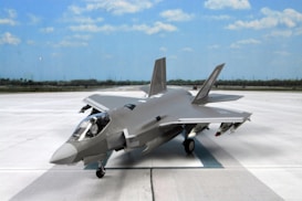 A modern military jet fighter is positioned on an expansive runway under a bright blue sky. The aircraft features sleek, aerodynamic lines and is painted in a gray camouflage pattern. It is equipped with various weapons and has a glossy cockpit canopy.