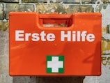 A bright orange first aid box mounted against a textured brick wall. The box features the words 'Erste Hilfe' in bold white letters, along with a green cross symbol, indicating first aid supplies.