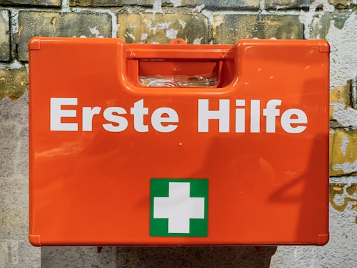 A bright orange first aid box mounted against a textured brick wall. The box features the words 'Erste Hilfe' in bold white letters, along with a green cross symbol, indicating first aid supplies.