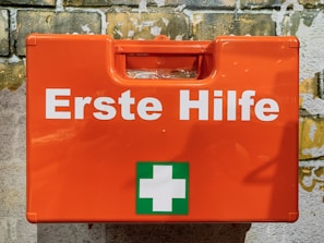 A bright orange first aid box mounted against a textured brick wall. The box features the words 'Erste Hilfe' in bold white letters, along with a green cross symbol, indicating first aid supplies.