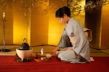 A person wearing a traditional Japanese kimono is performing a tea ceremony. The setting includes a tatami mat, a tea kettle, and a tray with ceremonial items. The room has a serene atmosphere with a warm yellow backdrop and cherry blossom branches overhead.