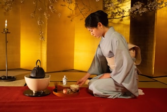 A person wearing a traditional Japanese kimono is performing a tea ceremony. The setting includes a tatami mat, a tea kettle, and a tray with ceremonial items. The room has a serene atmosphere with a warm yellow backdrop and cherry blossom branches overhead.
