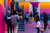A vibrant outdoor basketball court filled with people of various ages engaged in different activities. Some are playing basketball while others are standing and chatting. The court is surrounded by brightly painted walls in shades of pink, orange, and blue. A basketball hoop is mounted on the back wall, adding to the sports theme. A few people are sitting on stairs or using phones, creating a relaxed and social atmosphere.