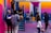 A vibrant outdoor basketball court filled with people of various ages engaged in different activities. Some are playing basketball while others are standing and chatting. The court is surrounded by brightly painted walls in shades of pink, orange, and blue. A basketball hoop is mounted on the back wall, adding to the sports theme. A few people are sitting on stairs or using phones, creating a relaxed and social atmosphere.