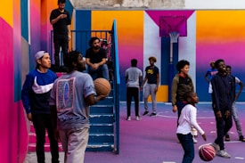 A vibrant outdoor basketball court filled with people of various ages engaged in different activities. Some are playing basketball while others are standing and chatting. The court is surrounded by brightly painted walls in shades of pink, orange, and blue. A basketball hoop is mounted on the back wall, adding to the sports theme. A few people are sitting on stairs or using phones, creating a relaxed and social atmosphere.