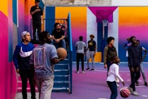 A vibrant outdoor basketball court filled with people of various ages engaged in different activities. Some are playing basketball while others are standing and chatting. The court is surrounded by brightly painted walls in shades of pink, orange, and blue. A basketball hoop is mounted on the back wall, adding to the sports theme. A few people are sitting on stairs or using phones, creating a relaxed and social atmosphere.
