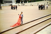 A couple wearing traditional, colorful clothing with intricate patterns stands on a set of steps in an open plaza. The woman wears a long, flowing red dress with a white headscarf, while the man is dressed in a striped robe. In the background, several groups of people are scattered across the wide, paved area.