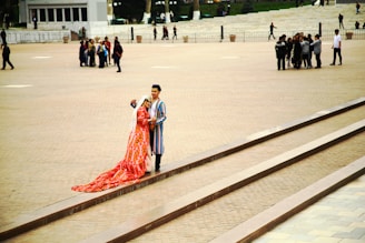 A couple wearing traditional, colorful clothing with intricate patterns stands on a set of steps in an open plaza. The woman wears a long, flowing red dress with a white headscarf, while the man is dressed in a striped robe. In the background, several groups of people are scattered across the wide, paved area.