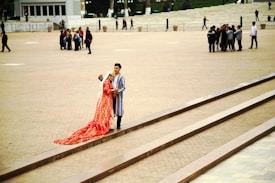A couple wearing traditional, colorful clothing with intricate patterns stands on a set of steps in an open plaza. The woman wears a long, flowing red dress with a white headscarf, while the man is dressed in a striped robe. In the background, several groups of people are scattered across the wide, paved area.