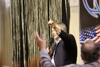 A man is speaking into a microphone, gesturing with one finger raised. He is addressing an audience, with raised hands visible. An American flag and a logo are partially visible in the background, alongside shimmering vertical strands.