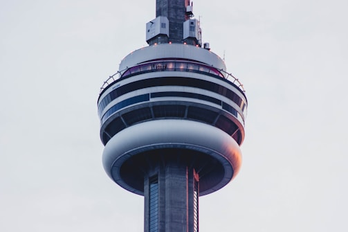 A close-up view of the upper section of a large telecommunications tower, featuring circular observation decks and various antennae. The structure is predominantly made of concrete and metal, with a sleek, modern design. The lighting appears soft due to the overcast sky.