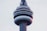 A close-up view of the upper section of a large telecommunications tower, featuring circular observation decks and various antennae. The structure is predominantly made of concrete and metal, with a sleek, modern design. The lighting appears soft due to the overcast sky.