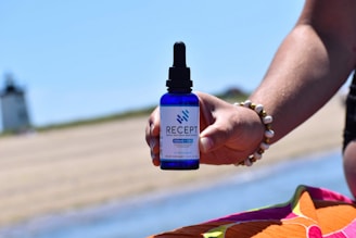 A hand is holding a blue dropper bottle labeled 'Recept' against a beach background. The bottle seems to contain broad spectrum hemp extract with CBG and CBC. The person is wearing a beaded bracelet, and there is a colorful fabric in the foreground. In the distance, a lighthouse is visible.