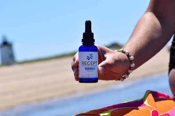 A hand is holding a blue dropper bottle labeled 'Recept' against a beach background. The bottle seems to contain broad spectrum hemp extract with CBG and CBC. The person is wearing a beaded bracelet, and there is a colorful fabric in the foreground. In the distance, a lighthouse is visible.