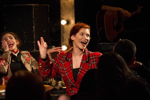 Two women are engaged in animated conversation or performance in a dimly lit setting, with a guitar player partially visible in the background. The women are wearing patterned jackets, and one has a microphone on her cheek. They appear to be lively and expressive.