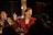Two women are engaged in animated conversation or performance in a dimly lit setting, with a guitar player partially visible in the background. The women are wearing patterned jackets, and one has a microphone on her cheek. They appear to be lively and expressive.