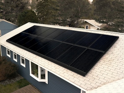 A set of black solar panels installed on the roof of a blue house with white window frames. The rooftop is covered with light gray shingles. The background includes several trees and another house in the distance.