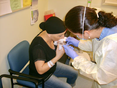 A healthcare professional is assisting a patient in a clinical setting. The patient is sitting on a chair, holding a cup near her mouth. The healthcare worker is wearing gloves and a protective gown, attending to the patient with what appears to be a medical procedure or care routine.