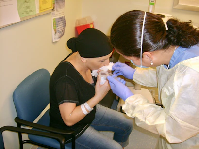 A healthcare professional is assisting a patient in a clinical setting. The patient is sitting on a chair, holding a cup near her mouth. The healthcare worker is wearing gloves and a protective gown, attending to the patient with what appears to be a medical procedure or care routine.