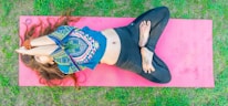 A person with long hair is lying on a pink yoga mat outdoors. They are in a yoga pose, with legs crossed and feet bare, wearing patterned clothing and a beaded bracelet. The grass surrounds the mat, adding a natural background.