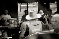 A monochrome photograph capturing a busy street scene. A person wearing a wide-brimmed hat faces away from the camera, gesturing with their hand while engaging with other individuals who seem to be focused on an activity. To the left, a sign with Thai text is partially visible. The setting appears to be an outdoor market or a bustling urban environment.