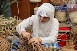 A person wearing traditional clothing is weaving with natural materials, surrounded by woven baskets and textiles. The setting appears to be a craft or artisan environment with handmade items and a natural aesthetic.