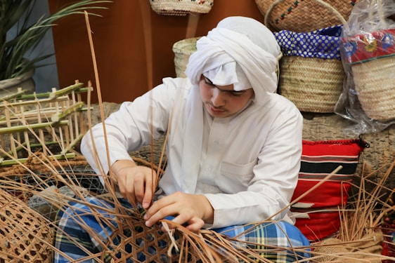 A person wearing traditional clothing is weaving with natural materials, surrounded by woven baskets and textiles. The setting appears to be a craft or artisan environment with handmade items and a natural aesthetic.