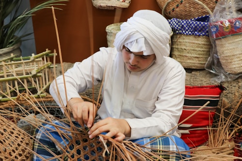 A person wearing traditional clothing is weaving with natural materials, surrounded by woven baskets and textiles. The setting appears to be a craft or artisan environment with handmade items and a natural aesthetic.