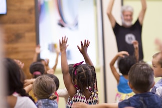 A group of young children enthusiastically raise their hands during an activity, with a bearded adult standing in the background participating as well. The setting appears to be a classroom or educational environment with colorful walls.