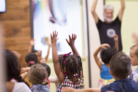A group of young children enthusiastically raise their hands during an activity, with a bearded adult standing in the background participating as well. The setting appears to be a classroom or educational environment with colorful walls.