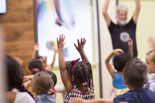 A group of young children enthusiastically raise their hands during an activity, with a bearded adult standing in the background participating as well. The setting appears to be a classroom or educational environment with colorful walls.