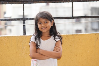 A young girl with long dark hair stands with her arms folded, smiling warmly. She is wearing a white t-shirt and is positioned against a bright yellow wall. The background includes a black metal railing and an out-of-focus urban setting.