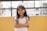 A young girl with long dark hair stands with her arms folded, smiling warmly. She is wearing a white t-shirt and is positioned against a bright yellow wall. The background includes a black metal railing and an out-of-focus urban setting.