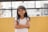 A young girl with long dark hair stands with her arms folded, smiling warmly. She is wearing a white t-shirt and is positioned against a bright yellow wall. The background includes a black metal railing and an out-of-focus urban setting.