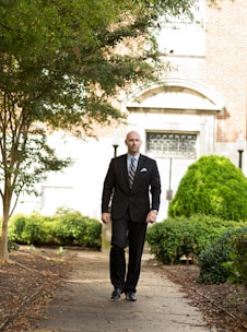 A man in a black suit walks down a path surrounded by lush greenery. The background features a building with a brick facade and decorative arched windows. The path is lined with trees and bushes, suggesting a tranquil outdoor setting.