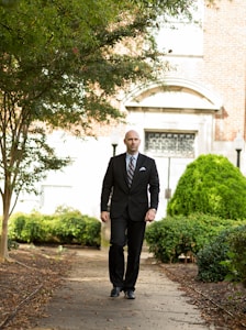 A man in a black suit walks down a path surrounded by lush greenery. The background features a building with a brick facade and decorative arched windows. The path is lined with trees and bushes, suggesting a tranquil outdoor setting.