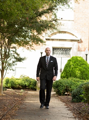 A man in a black suit walks down a path surrounded by lush greenery. The background features a building with a brick facade and decorative arched windows. The path is lined with trees and bushes, suggesting a tranquil outdoor setting.