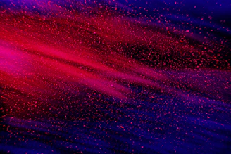 A dynamic blend of vibrant red and deep blue colors with a speckled, dust-like texture that conveys a sense of motion and energy.