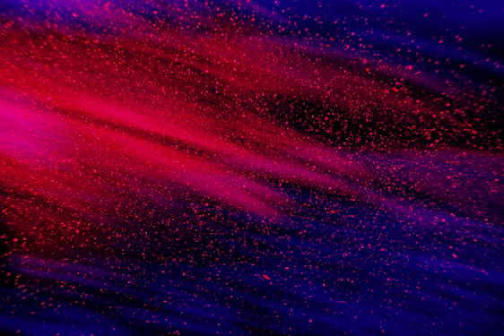 A dynamic blend of vibrant red and deep blue colors with a speckled, dust-like texture that conveys a sense of motion and energy.