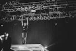 A performer stands on an elevated stage structure, reaching up towards a rigged lighting setup in a dimly lit environment. The atmosphere is dynamic and energetic, captured in black and white, with the focus on the interaction between the performer and the stage lights.