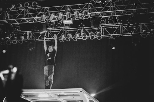 A performer stands on an elevated stage structure, reaching up towards a rigged lighting setup in a dimly lit environment. The atmosphere is dynamic and energetic, captured in black and white, with the focus on the interaction between the performer and the stage lights.